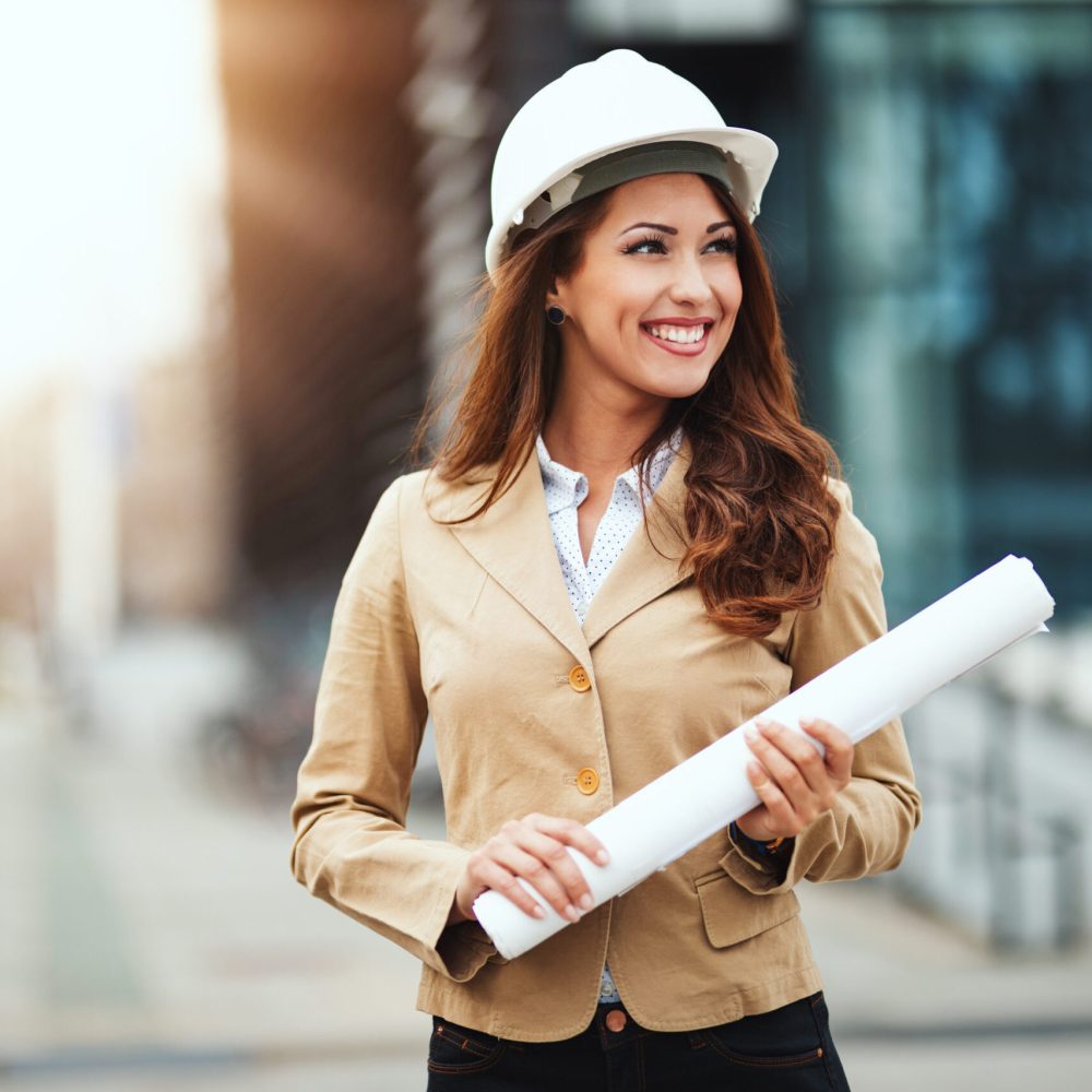 Portrait of young engineer woman with white helmet and blueprints in her hands at construction site.